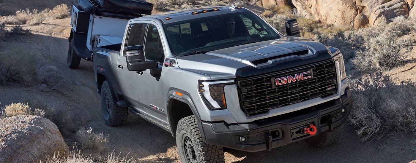 A silver 2026 GMC Sierra 2500 HD AT4X towing a trailer up rocky terrain.